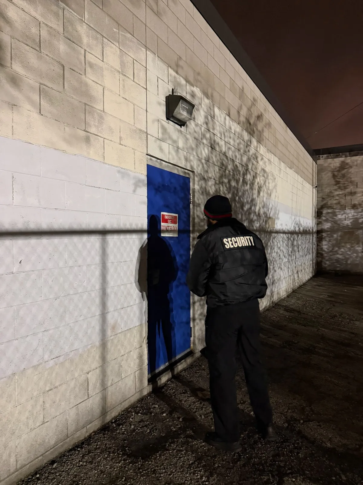 Security guard in a black jacket stands near a blue emergency exit door on a white brick wall at night.